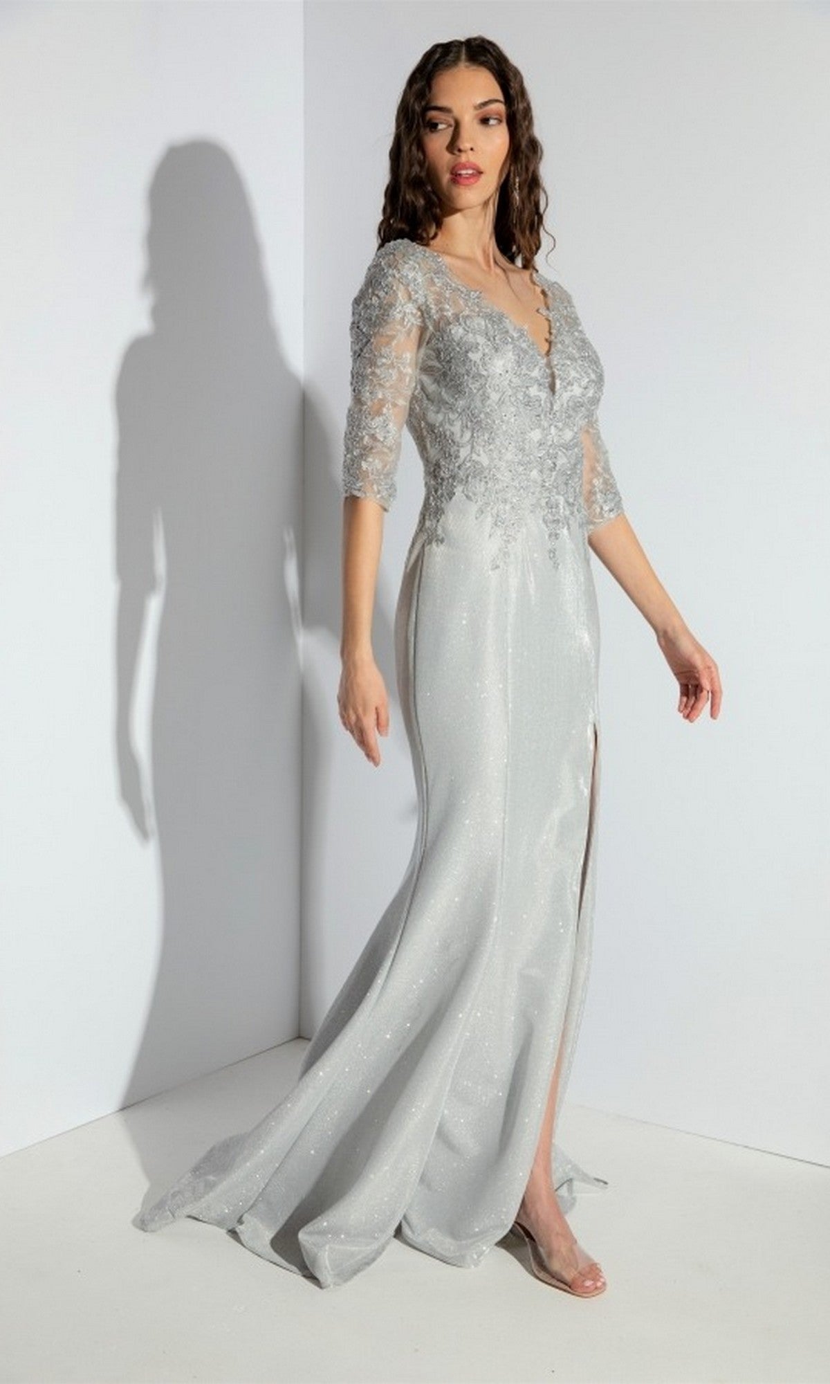 A woman stands against a white wall wearing the Eureka 9970 Long Prom Dress in silver, featuring a floor-length silhouette, embroidered bodice, lace sleeves, and a front slit.